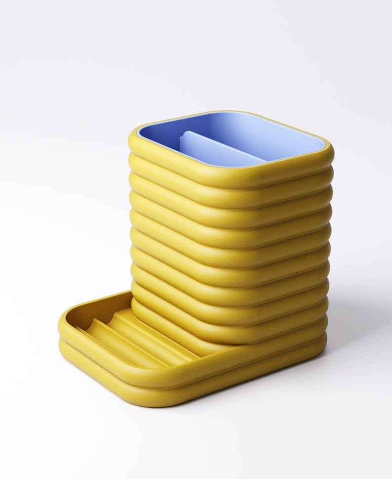 May include: A yellow desk organizer with a blue interior. The organizer has a rectangular shape with a series of horizontal ridges. A small tray is attached to the side. The design is modern and minimalist.