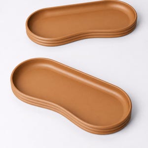 May include: Two brown, oval-shaped serving trays stacked on a white background. The trays have a smooth surface and a raised edge. The trays are a warm, earthy tone, perfect for serving food or displaying items.