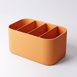 May include: Orange rectangular storage organiser with four compartments. The exterior has vertical ribbed detailing. The interior dividers are smooth. The organiser is made of a solid, opaque material, designed for storage and organisation.