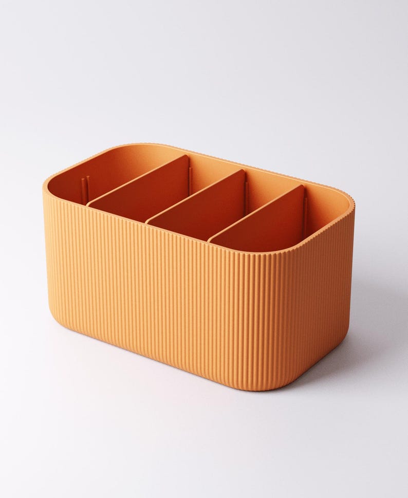 May include: Orange rectangular storage organiser with four compartments. The exterior has vertical ribbed detailing. The interior dividers are smooth. The organiser is made of a solid, opaque material, designed for storage and organisation.