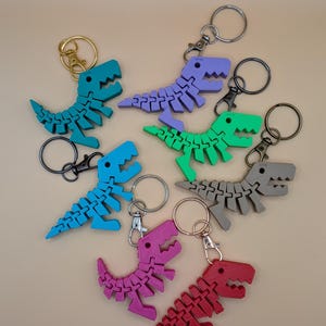 May include: A collection of colorful dinosaur keychains in various shades of blue, green, purple, pink, red, and gray. Each keychain features a segmented dinosaur design with a metal keyring and clasp. The keychains are arranged on a neutral background.