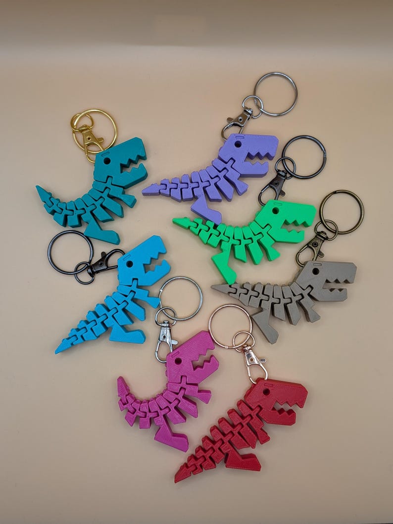 May include: A collection of colorful dinosaur keychains in various shades of blue, green, purple, pink, red, and gray. Each keychain features a segmented dinosaur design with a metal keyring and clasp. The keychains are arranged on a neutral background.