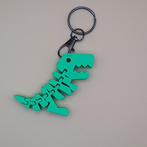 May include: A green articulated dinosaur keychain. The dinosaur skeleton is made of connected pieces, with a black metal clasp and keyring. The keychain is a fun accessory for bags, keys, or backpacks.