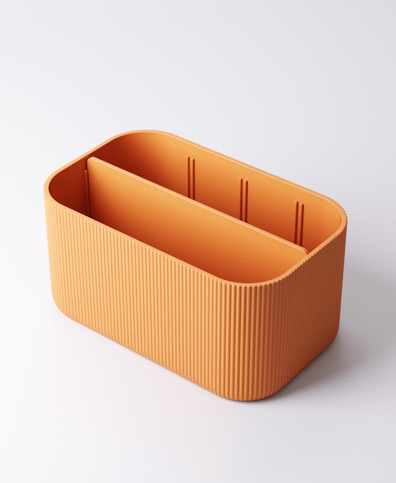 May include: Orange rectangular storage organiser with a ribbed exterior and two internal dividers. The organiser is made of a solid orange material and has a rounded shape. The dividers create three separate compartments.
