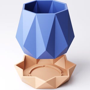 May include: A blue geometric planter and a peach-coloured saucer. The planter has a faceted design and a wide opening. The saucer has a star-like shape with a circular indentation. The planter and saucer are made of a solid material. The planter is approximately 15 cm tall.