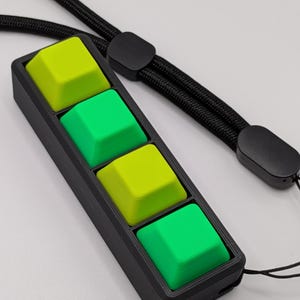 May include: A black rectangular device with four square, colorful buttons. Two buttons are bright green, and two are lime green. A black lanyard is attached to the device.