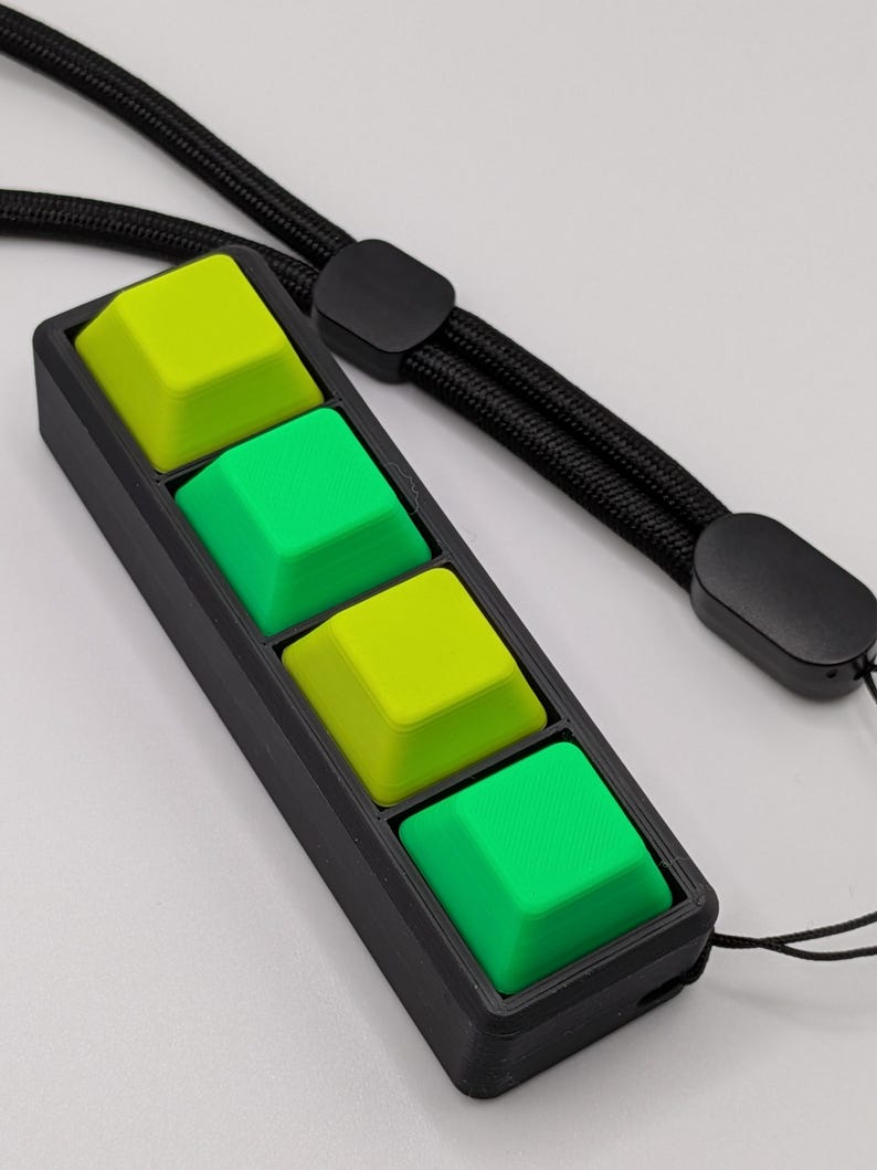 May include: A black rectangular device with four square, colorful buttons. Two buttons are bright green, and two are lime green. A black lanyard is attached to the device.