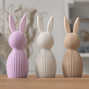 K&ouml;nnte beinhalten: Drei Holzhasenfiguren in Pastellfarben. Die Hasen haben ein vertikales Streifendesign und lange Ohren. Die Farben sind Lavendel, Wei&szlig; und Hellbraun. Diese dekorativen Hasen sind perfekt f&uuml;r Ostern.