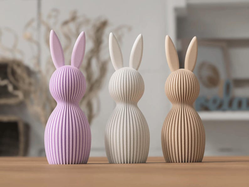 K&ouml;nnte beinhalten: Drei Holzhasenfiguren in Pastellfarben. Die Hasen haben ein vertikales Streifendesign und lange Ohren. Die Farben sind Lavendel, Wei&szlig; und Hellbraun. Diese dekorativen Hasen sind perfekt f&uuml;r Ostern.