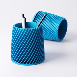K&ouml;nnte beinhalten: Zwei blaue Stifthalter mit Spiralmuster. Ein aufrechter Halter enth&auml;lt einen wei&szlig;en und einen schwarzen Stift. Der andere Halter liegt auf der Seite. Die Halter sind aus einem einfarbigen, hellblauen Material.