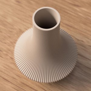 May include: A beige ceramic vase with a wide, ribbed base that tapers to a narrow neck. The vase has a circular opening at the top and is set against a wooden surface.