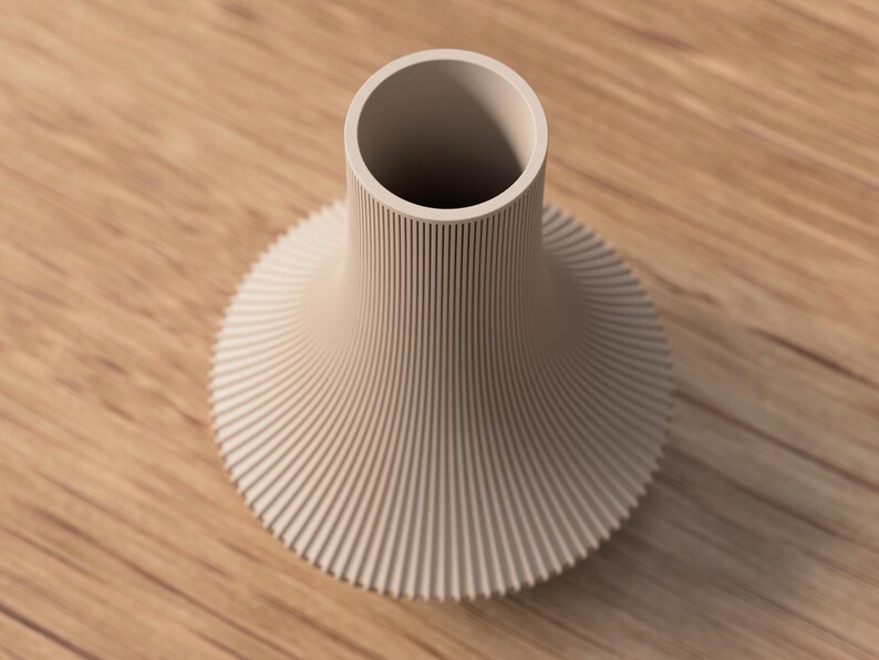 May include: A beige ceramic vase with a wide, ribbed base that tapers to a narrow neck. The vase has a circular opening at the top and is set against a wooden surface.