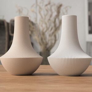 May include: Two decorative vases on a wooden surface. One vase is a textured beige, the other is white with vertical ribbed detailing. Both have a flared base and a narrow neck, suitable for home decor.
