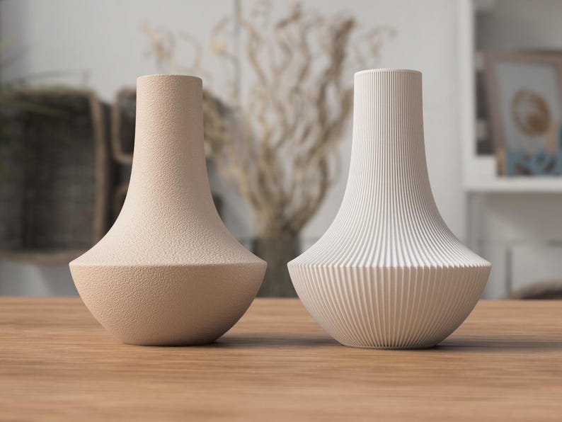 May include: Two decorative vases on a wooden surface. One vase is a textured beige, the other is white with vertical ribbed detailing. Both have a flared base and a narrow neck, suitable for home decor.
