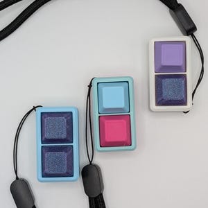 May include: Three keycap pendants in light blue, pink, and purple, each with a matching lanyard. The pendants have a two-key design, with a glittery, dark blue square below a solid-colored square. The lanyards are black.