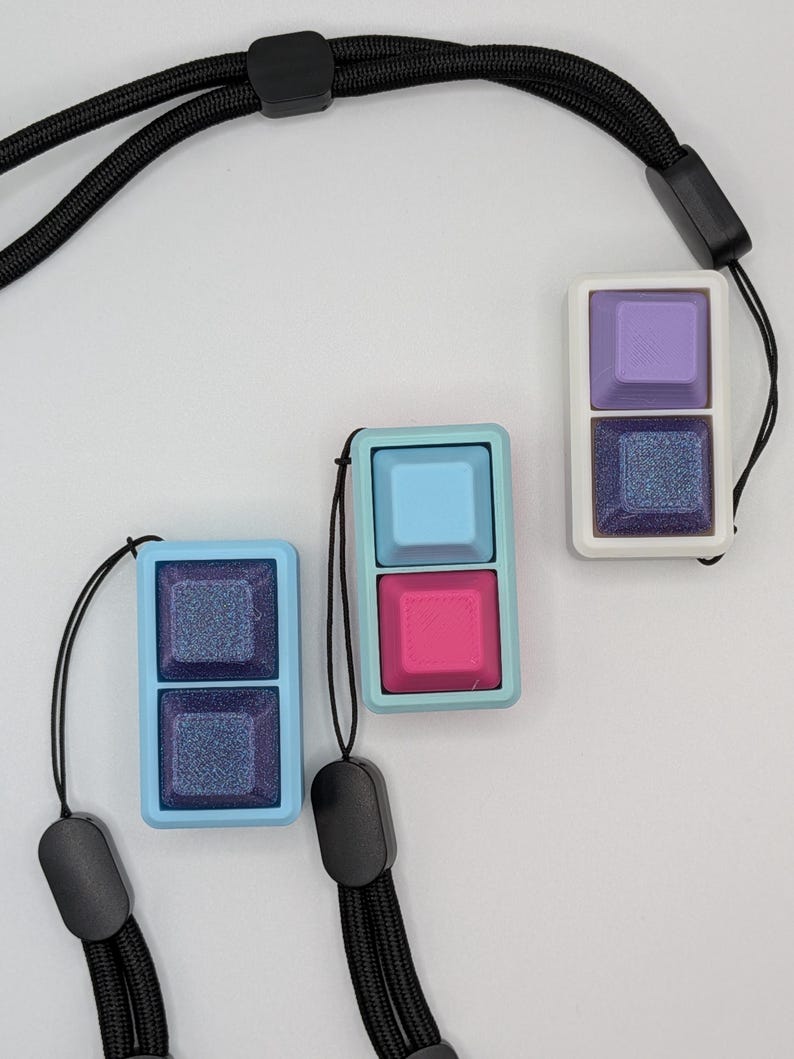 May include: Three keycap pendants in light blue, pink, and purple, each with a matching lanyard. The pendants have a two-key design, with a glittery, dark blue square below a solid-colored square. The lanyards are black.