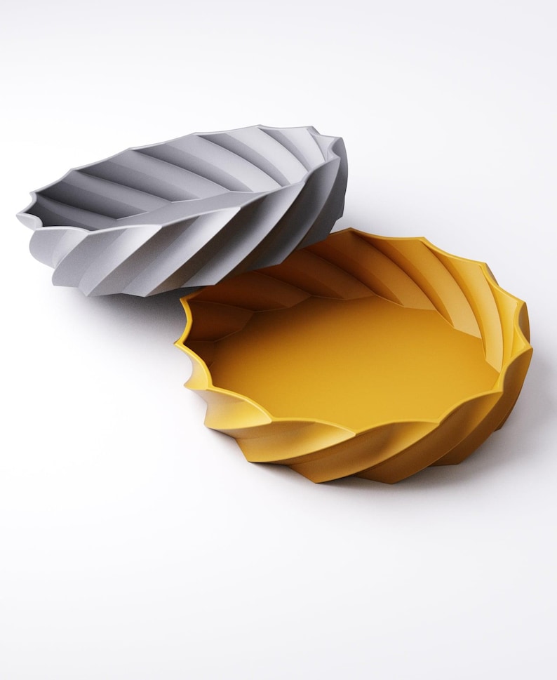 May include: Two decorative bowls with a unique spiral design. One bowl is a light gray color, and the other is a vibrant yellow. The bowls have a wavy, scalloped edge and are set against a white background. These bowls are ideal for home decor.