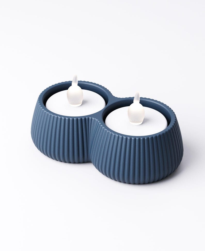 May include: A blue, double-vessel candle holder with a ribbed exterior. Each vessel holds a white disc with a small, white, flame-shaped light. The candle holder is a modern home decor item.