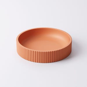 May include: A round, terracotta-colored bowl with a ribbed exterior. The bowl has a shallow interior and a textured, vertical ribbed design around the sides. The bowl is made of a solid material and is suitable for serving or display.
