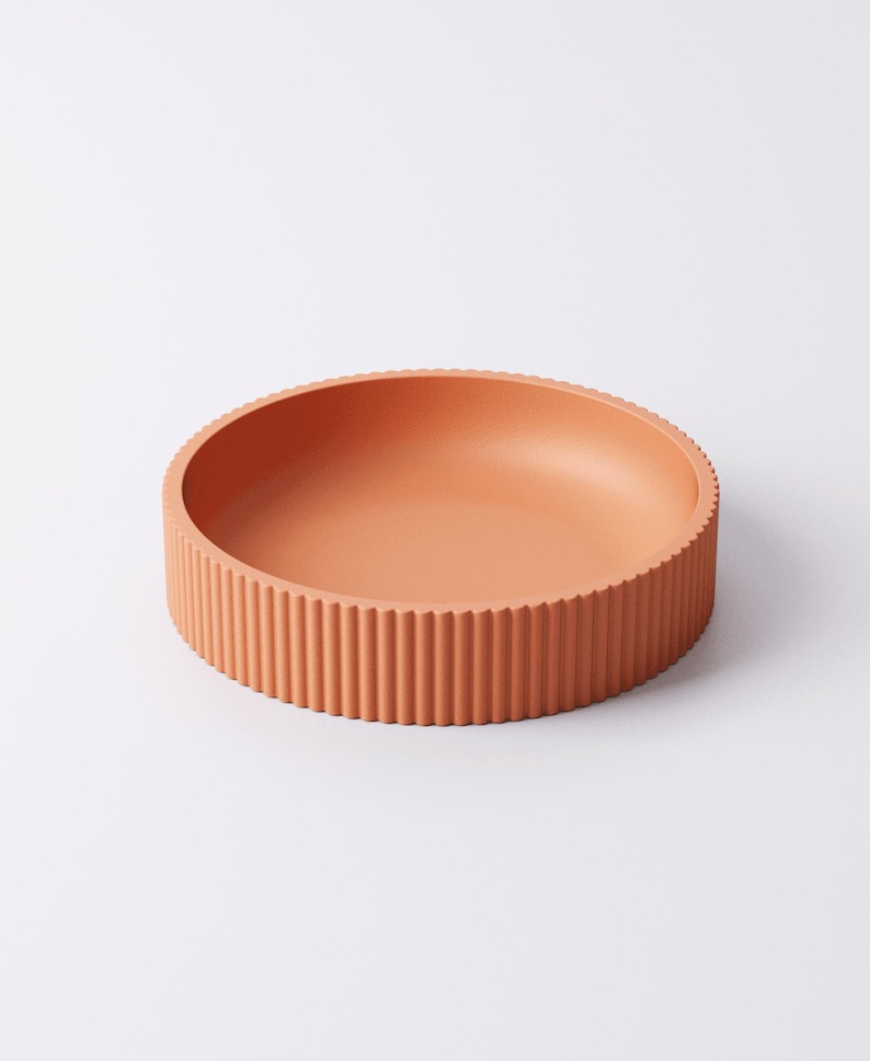 May include: A round, terracotta-colored bowl with a ribbed exterior. The bowl has a shallow interior and a textured, vertical ribbed design around the sides. The bowl is made of a solid material and is suitable for serving or display.
