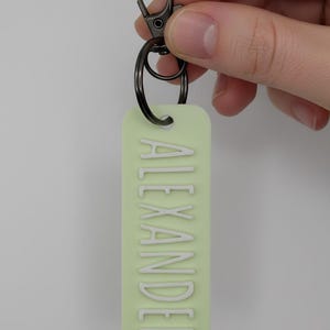 May include: A light green rectangular keychain with the name "ALEXANDER" in white capital letters. The keychain has a dark gray metal clasp and ring for attaching keys. The background is a solid light gray.