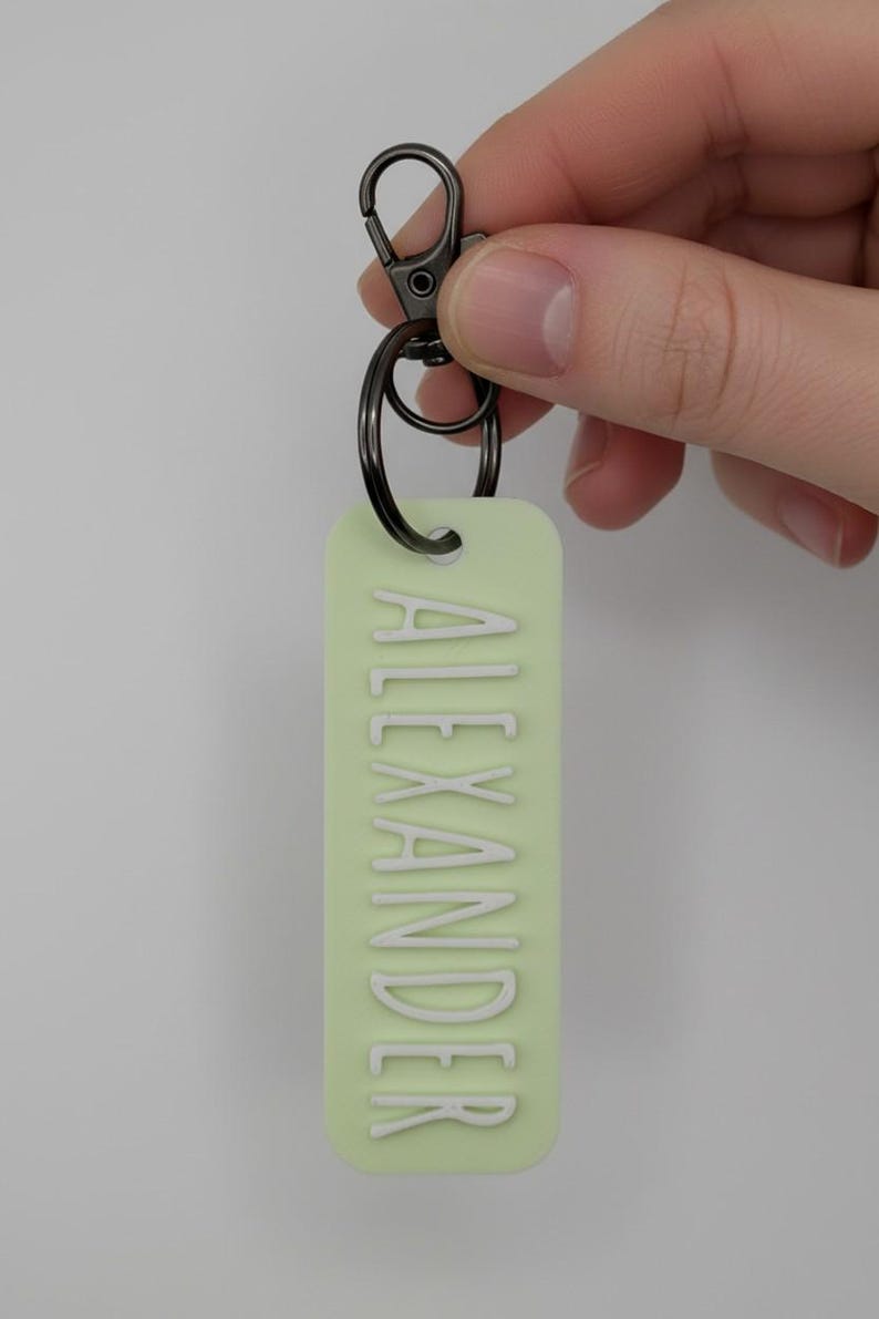 May include: A light green rectangular keychain with the name "ALEXANDER" in white capital letters. The keychain has a dark gray metal clasp and ring for attaching keys. The background is a solid light gray.