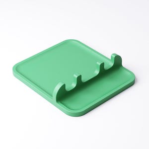 May include: A square, light green spoon rest with three curved slots for holding utensils. The spoon rest is made of a solid material and has a slightly raised edge. The design is simple and modern, suitable for kitchen use.