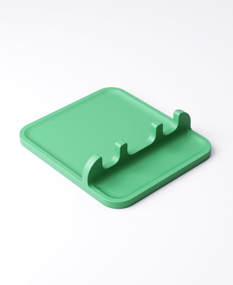 May include: A square, light green spoon rest with three curved slots for holding utensils. The spoon rest is made of a solid material and has a slightly raised edge. The design is simple and modern, suitable for kitchen use.