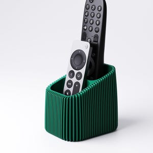 May include: A green ribbed remote control holder with two remotes. One remote is black with white numbers and buttons, and the other is silver with black buttons. The holder is a solid, dark green color.