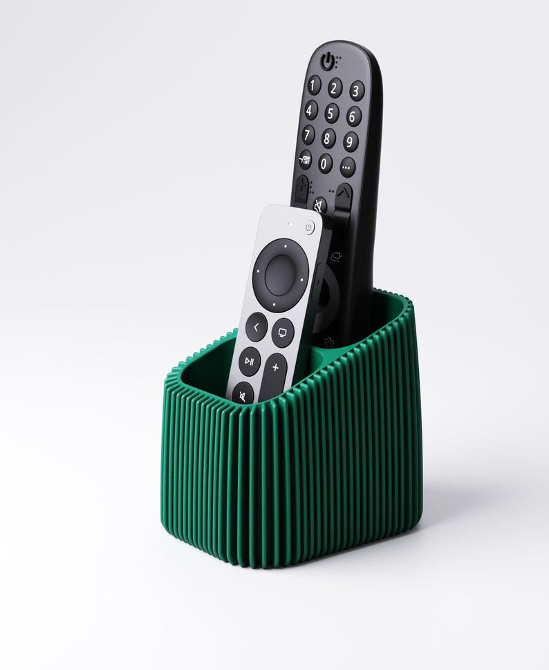 May include: A green ribbed remote control holder with two remotes. One remote is black with white numbers and buttons, and the other is silver with black buttons. The holder is a solid, dark green color.