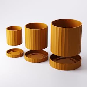 Puede incluir: Tres macetas cil&iacute;ndricas de color amarillo mostaza de diferentes tama&ntilde;os, cada una con una textura acanalada vertical en el exterior. Cada maceta viene acompa&ntilde;ada de un plato circular a juego con un borde acanalado.