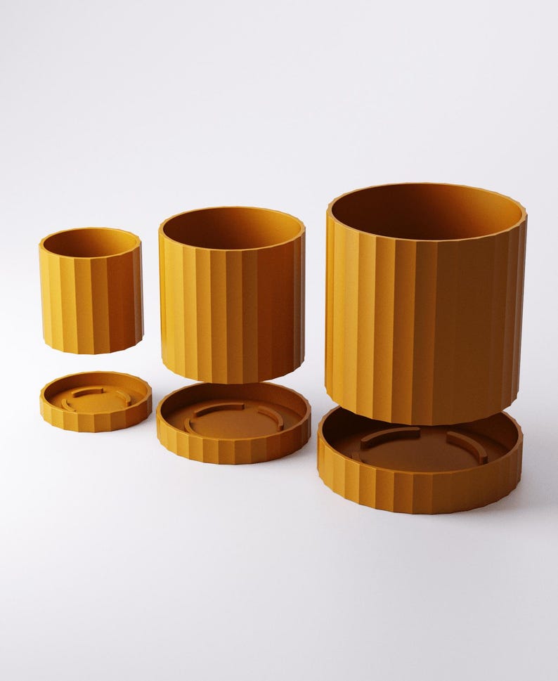 Puede incluir: Tres macetas cil&iacute;ndricas de color amarillo mostaza de diferentes tama&ntilde;os, cada una con una textura acanalada vertical en el exterior. Cada maceta viene acompa&ntilde;ada de un plato circular a juego con un borde acanalado.