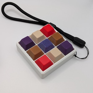 May include: A square, white plastic case holds nine multi-colored keycaps. The keycaps are red, tan, brown, purple, and blue. A black lanyard is attached to the case. The keycaps are arranged in a 3x3 grid.