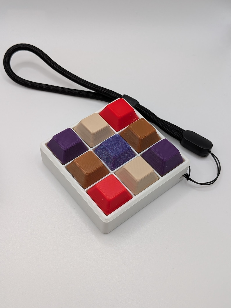May include: A square, white plastic case holds nine multi-colored keycaps. The keycaps are red, tan, brown, purple, and blue. A black lanyard is attached to the case. The keycaps are arranged in a 3x3 grid.