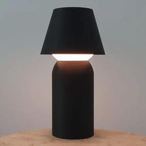 May include: A modern black table lamp with a cylindrical base and a lampshade that tapers upwards. The lamp has a bright, horizontal light source. The lamp is on a wooden surface.