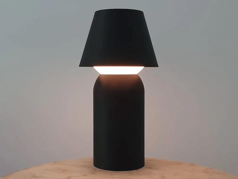 May include: A modern black table lamp with a cylindrical base and a lampshade that tapers upwards. The lamp has a bright, horizontal light source. The lamp is on a wooden surface.