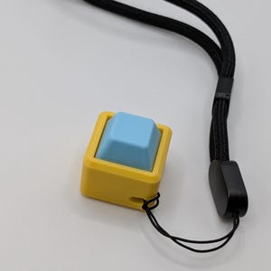 May include: A yellow and blue keycap with a black wrist strap. The keycap has a square shape with a light blue top and a yellow frame. The black wrist strap is made of a woven material.