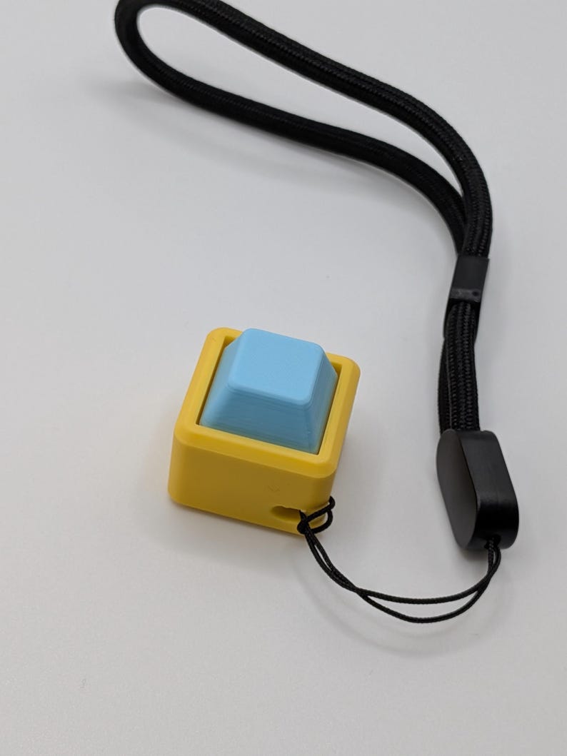 May include: A yellow and blue keycap with a black wrist strap. The keycap has a square shape with a light blue top and a yellow frame. The black wrist strap is made of a woven material.