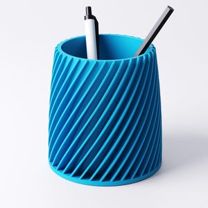 K&ouml;nnte beinhalten: Ein leuchtend blauer Stifthalter mit einem Spiralmuster. Er enth&auml;lt einen wei&szlig;-schwarzen Stift und einen schwarz-grauen Stift. Der Stifthalter besteht aus einem festen Material und steht auf einer wei&szlig;en Oberfl&auml;che.