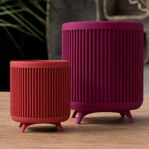 Puede incluir: Dos macetas cil&iacute;ndricas, una roja y otra magenta, sobre una superficie de madera. Ambas tienen detalles verticales acanalados y est&aacute;n soportadas por tres peque&ntilde;as patas redondeadas. Las macetas est&aacute;n vac&iacute;as.