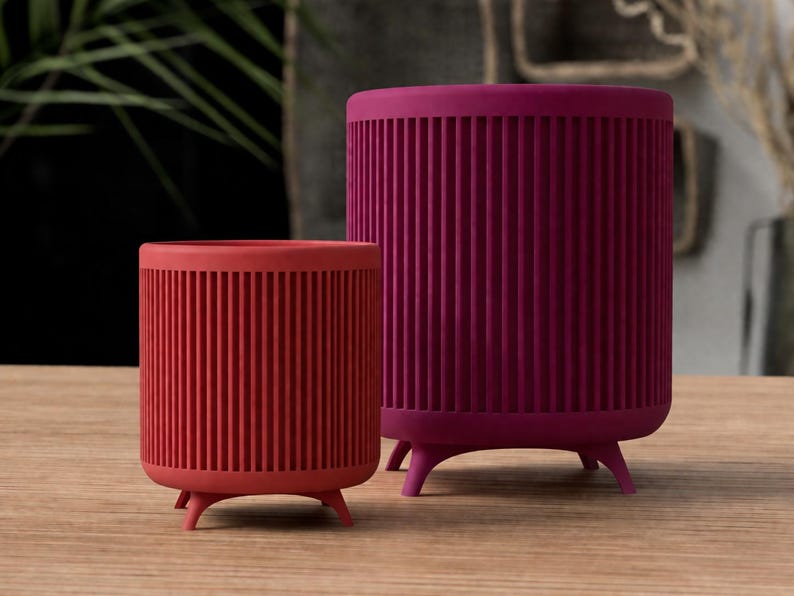 Puede incluir: Dos macetas cil&iacute;ndricas, una roja y otra magenta, sobre una superficie de madera. Ambas tienen detalles verticales acanalados y est&aacute;n soportadas por tres peque&ntilde;as patas redondeadas. Las macetas est&aacute;n vac&iacute;as.
