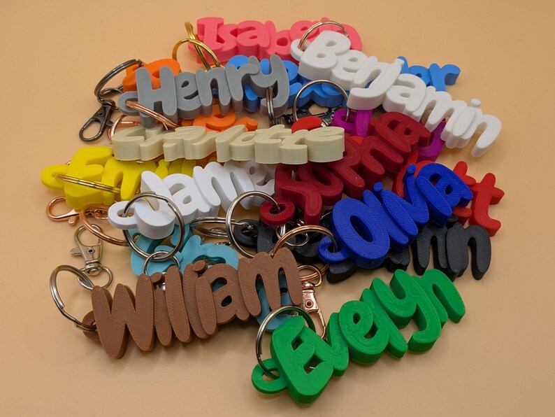 May include: A collection of colorful name keychains in various colors, including pink, blue, green, and brown. Each keychain features a different name in a 3D font, attached to a metal keyring. The keychains are scattered on a light orange surface.