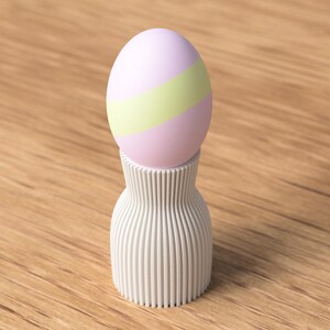 Pu&ograve; includere: Un uovo di Pasqua rosa pastello e verde &egrave; appoggiato su un portauovo bianco a coste verticali. L'uovo ha una striscia verde chiaro al centro. Il portauovo ha la forma di un vaso. Lo sfondo &egrave; una superficie di legno marrone chiaro.