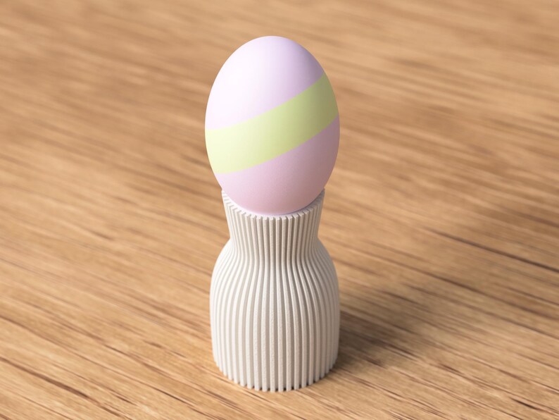 Pu&ograve; includere: Un uovo di Pasqua rosa pastello e verde &egrave; appoggiato su un portauovo bianco a coste verticali. L'uovo ha una striscia verde chiaro al centro. Il portauovo ha la forma di un vaso. Lo sfondo &egrave; una superficie di legno marrone chiaro.