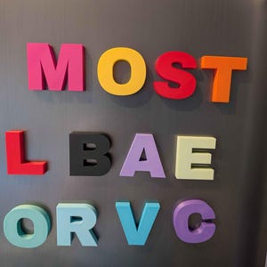 May include: Colorful magnetic letters spelling out "MOST L BAE OR VC" on a gray refrigerator. The letters are in various colors, including pink, yellow, red, orange, black, lavender, light green, teal, and purple. The letters are arranged in three rows.