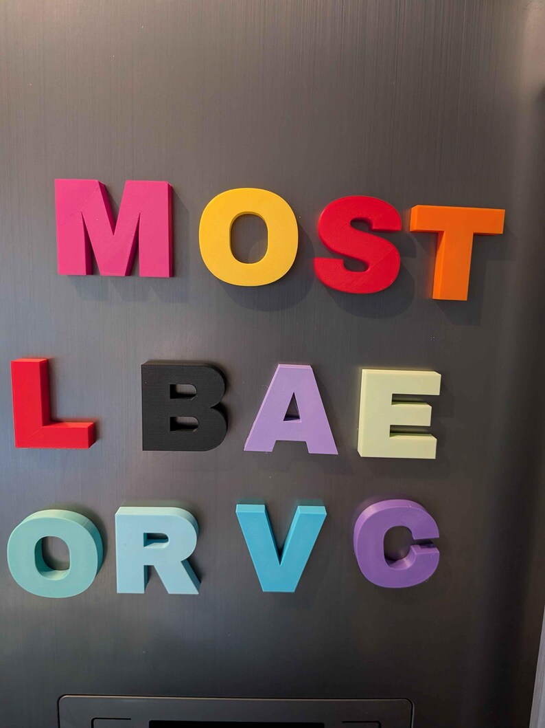 May include: Colorful magnetic letters spelling out "MOST L BAE OR VC" on a gray refrigerator. The letters are in various colors, including pink, yellow, red, orange, black, lavender, light green, teal, and purple. The letters are arranged in three rows.