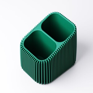 May include: A green, two-compartment desk organizer with a ribbed exterior. The organizer has a rectangular shape with rounded corners and a textured, vertical ribbed design. The interior compartments are smooth and open.