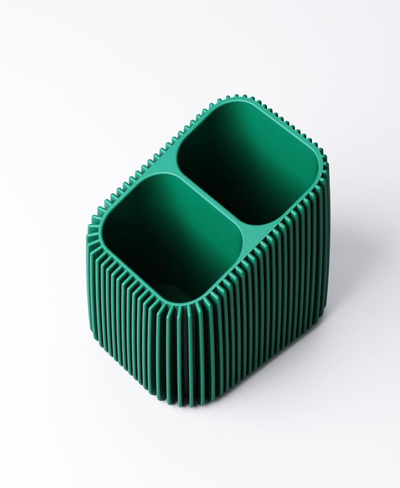 May include: A green, two-compartment desk organizer with a ribbed exterior. The organizer has a rectangular shape with rounded corners and a textured, vertical ribbed design. The interior compartments are smooth and open.