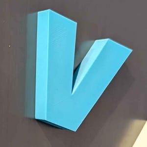 May include: A light blue, three-dimensional letter "V" made of a solid material. The letter is mounted on a dark gray surface. The "V" shape is angular and modern, with clean lines and a matte finish.