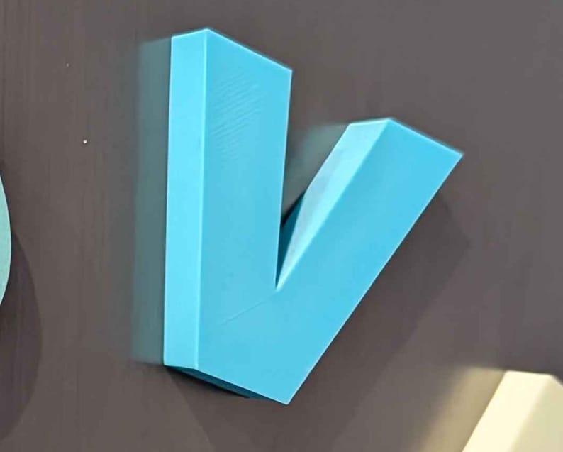 May include: A light blue, three-dimensional letter "V" made of a solid material. The letter is mounted on a dark gray surface. The "V" shape is angular and modern, with clean lines and a matte finish.